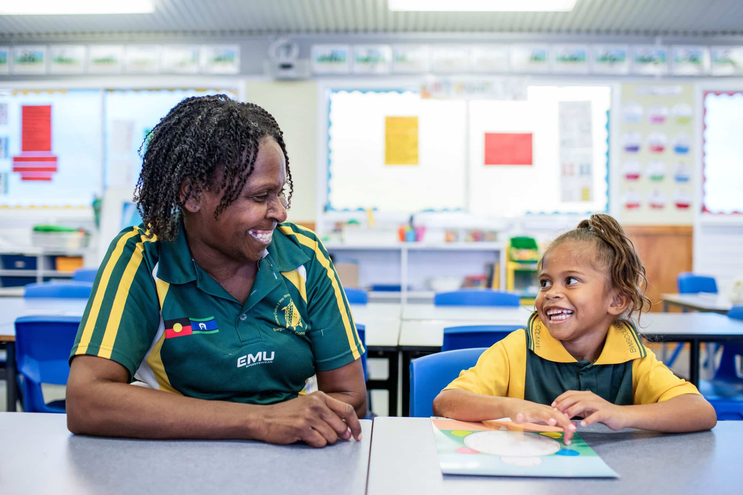 An adult and child sitting at desks in a classroom smiling at each other