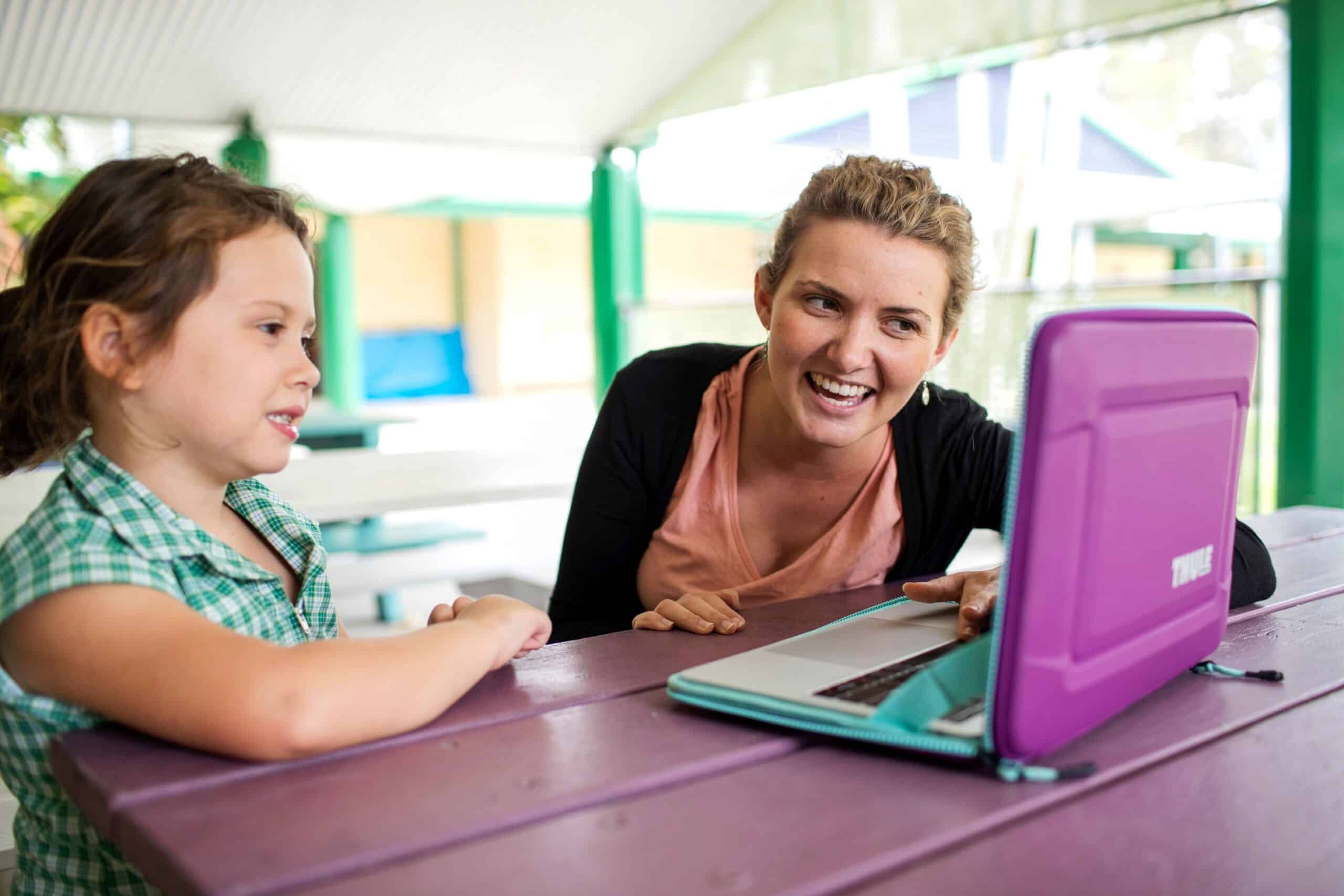 A child and adult sitting at a table, smiling and laughing while looking at a laptop